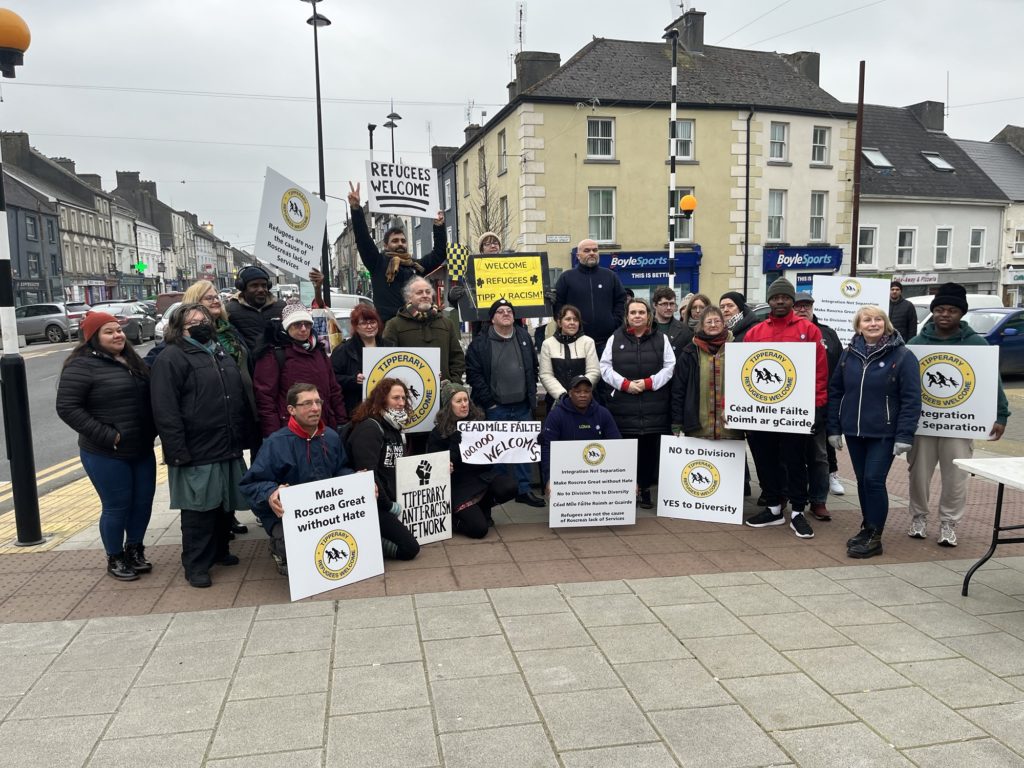 A group of people standing at the top of Castle Street in Roscrea holding placards saying “Refugees Welcome”, “Make Roscrea Great Without Hate”, “Céad Míle Fáilte Roimh Ár gCáirde” and others. 